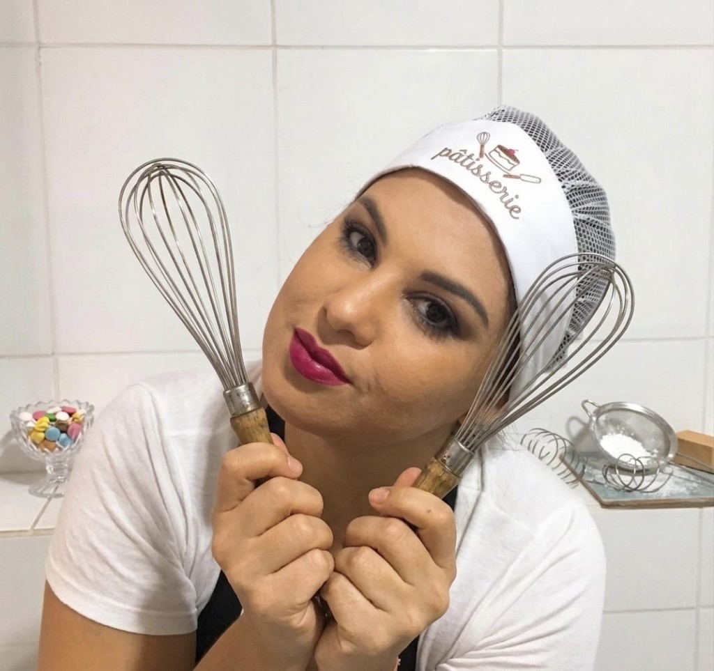 Alice, confeiteira da Doce Alice, com touca de pâtisserie e batedores, em cozinha com macarons ao fundo.